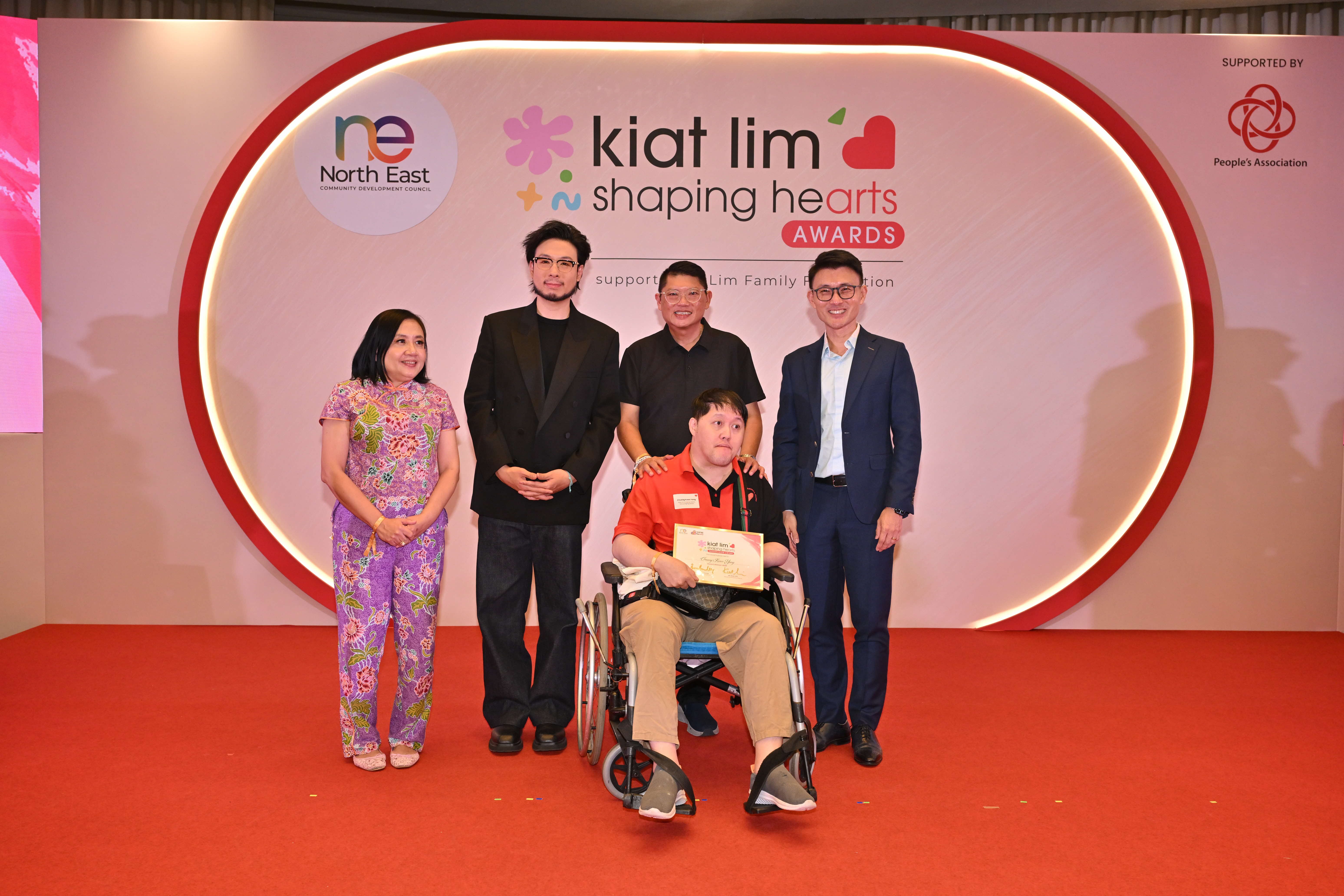 Award recipient standing on stage with the presenter, holding their certificate and posing for a group photo during the award ceremony, with a backdrop reading ‘Kiat Lim-Shaping Hearts Awards'.